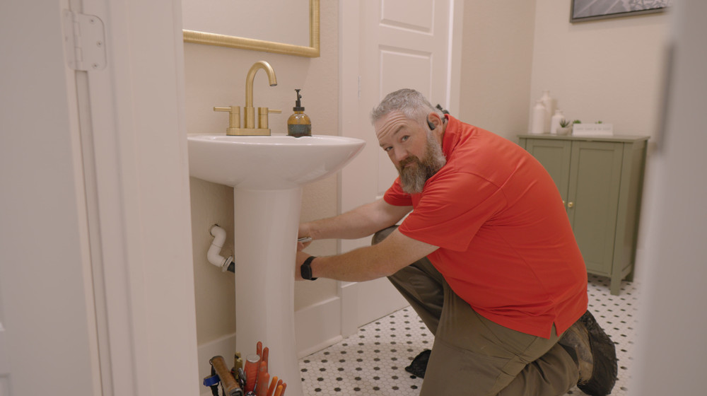 A plumber kneeling to work on the drain under a pedestal sink